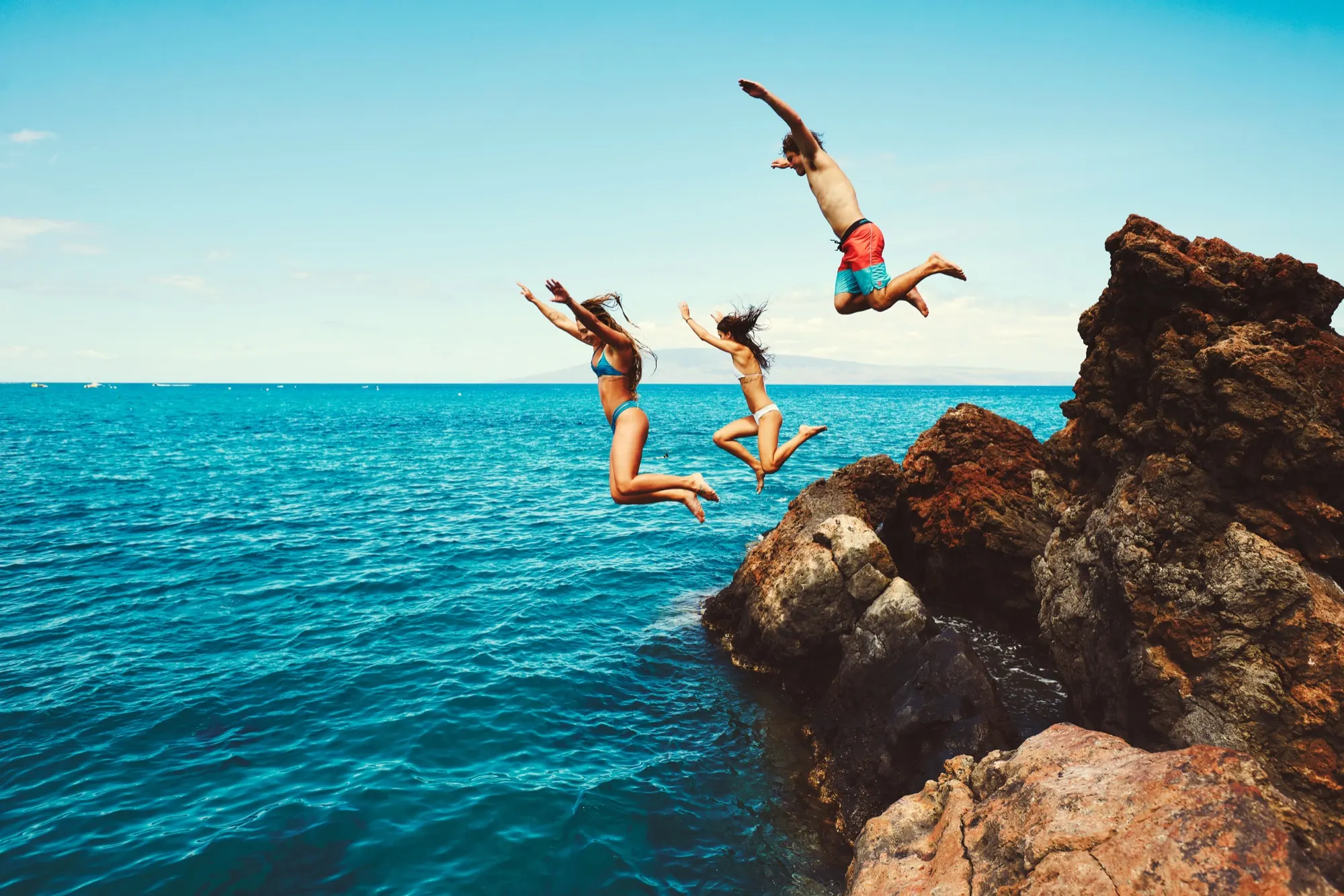 Friends leaping from a lava rock ledge into the turquoise Hawaiian ocean