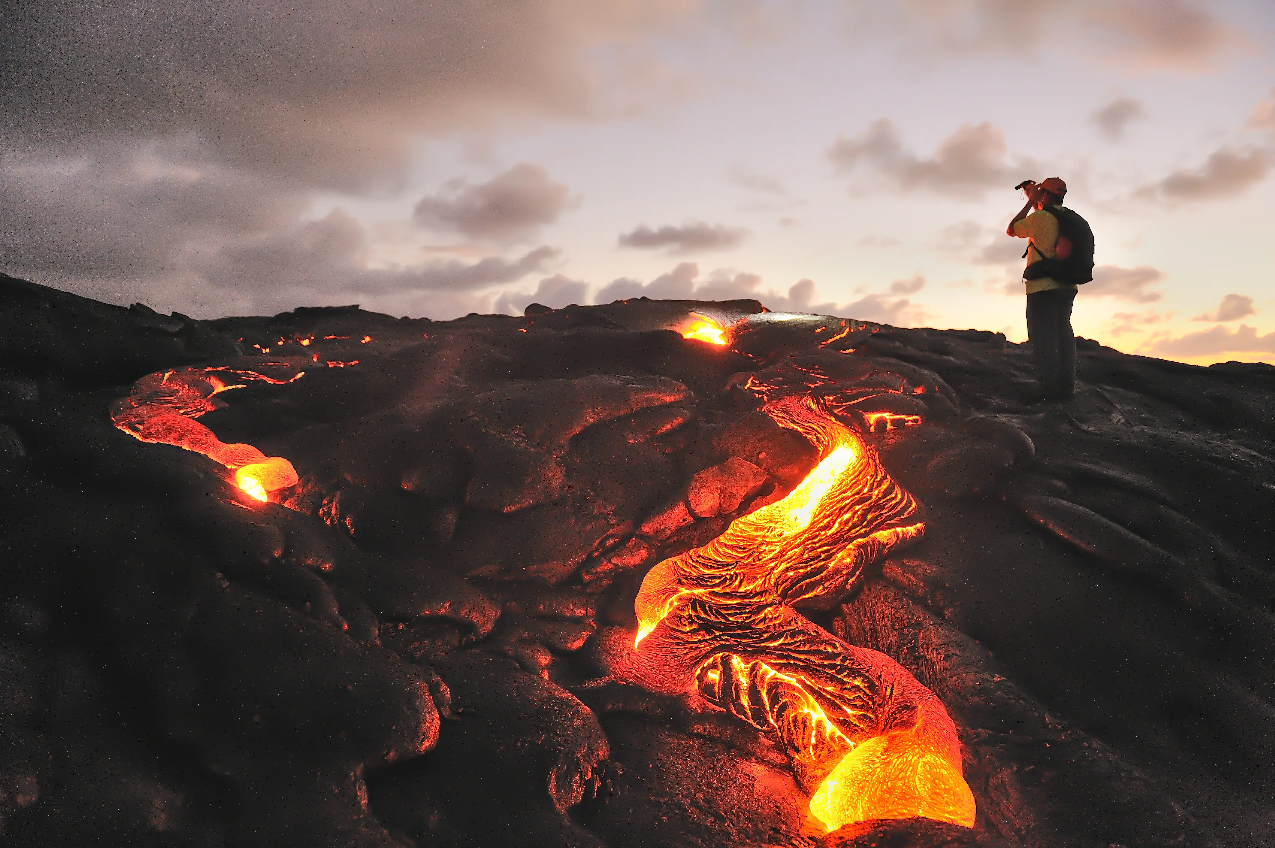 A lone traveler photographing a glowing river of lava at dusk on Hawaiʻi Island