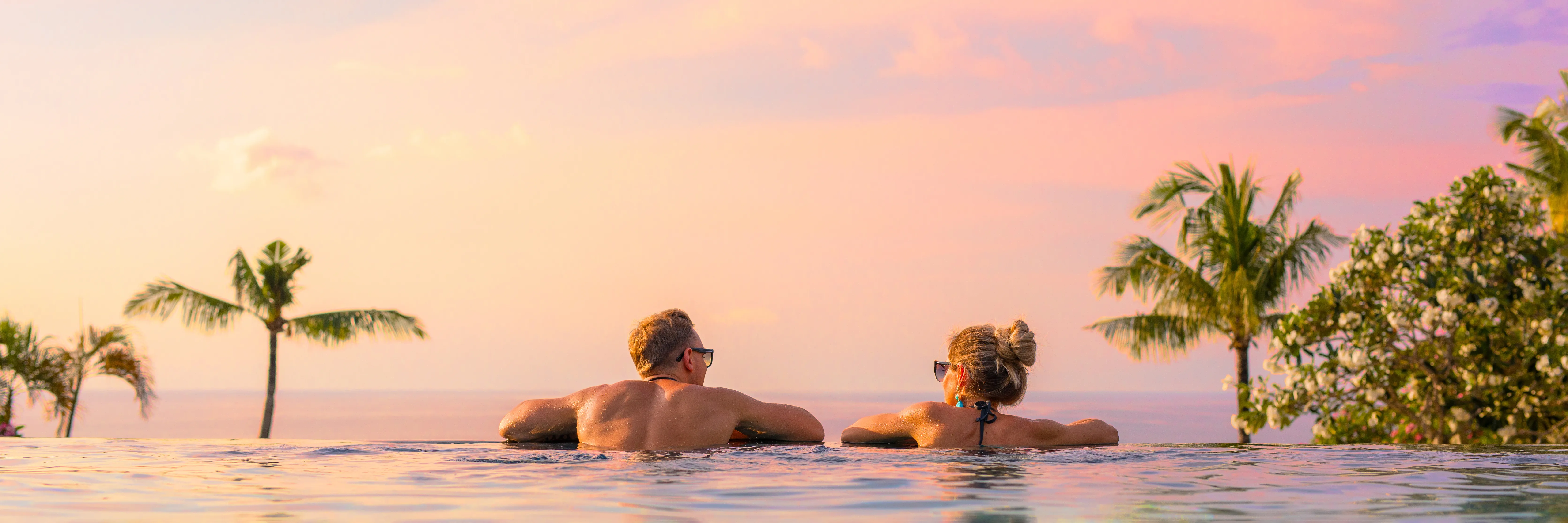 A couple relaxing in an infinity pool at sunset above the Hawaiʻi Island coastline