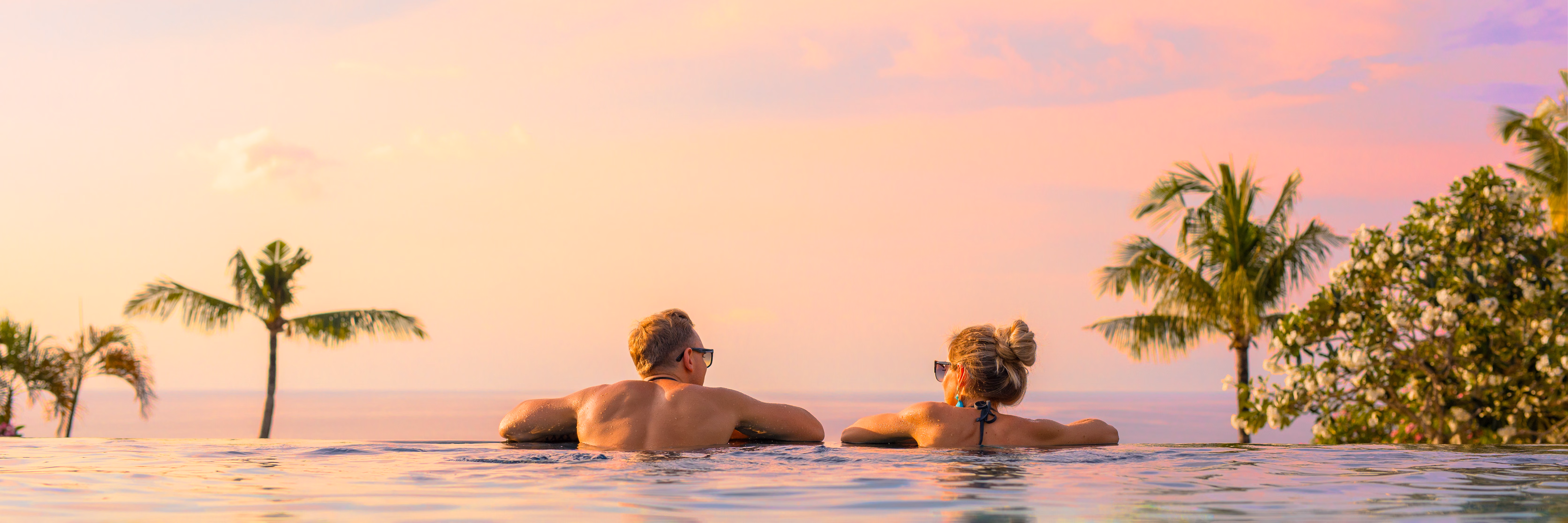A couple relaxing in an infinity pool at sunset above the Hawaiʻi Island coastline